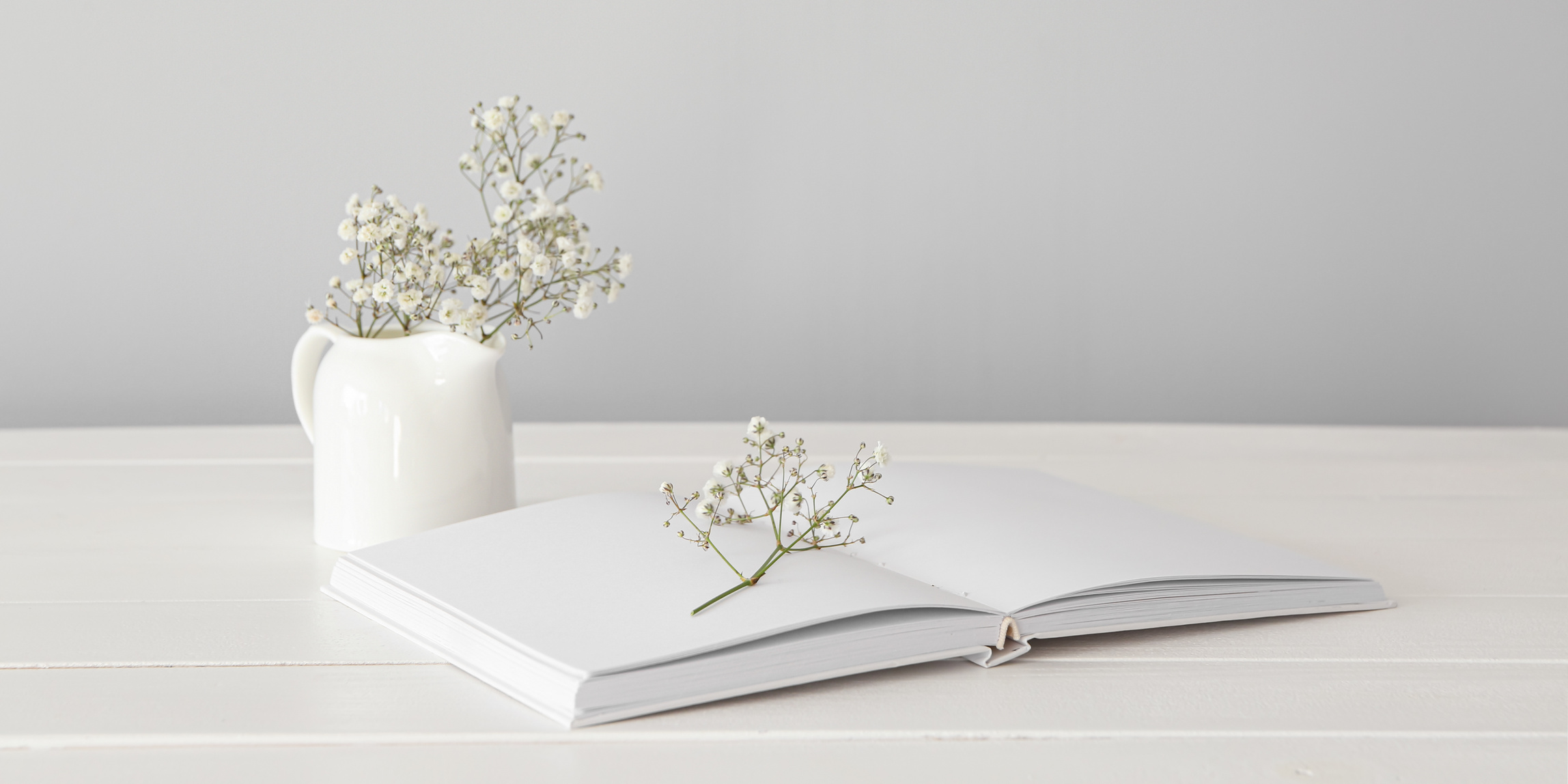 Open Book with Jug and Flowers on White Wooden Table against Light Background